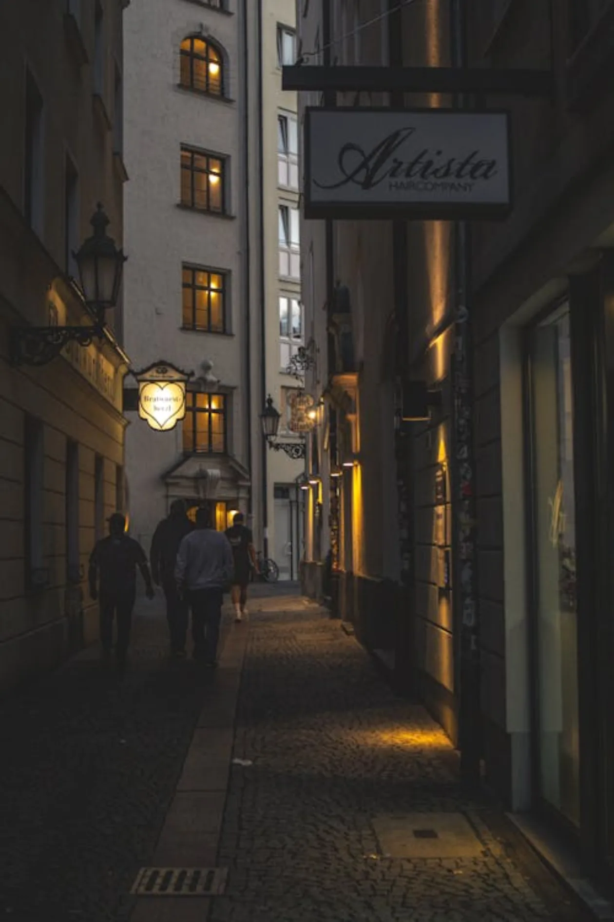 Moody urban alley at night with warm street lamps, long shadows, wet cobblestones, cinematic noir atmosphere, atmospheric urban texture
