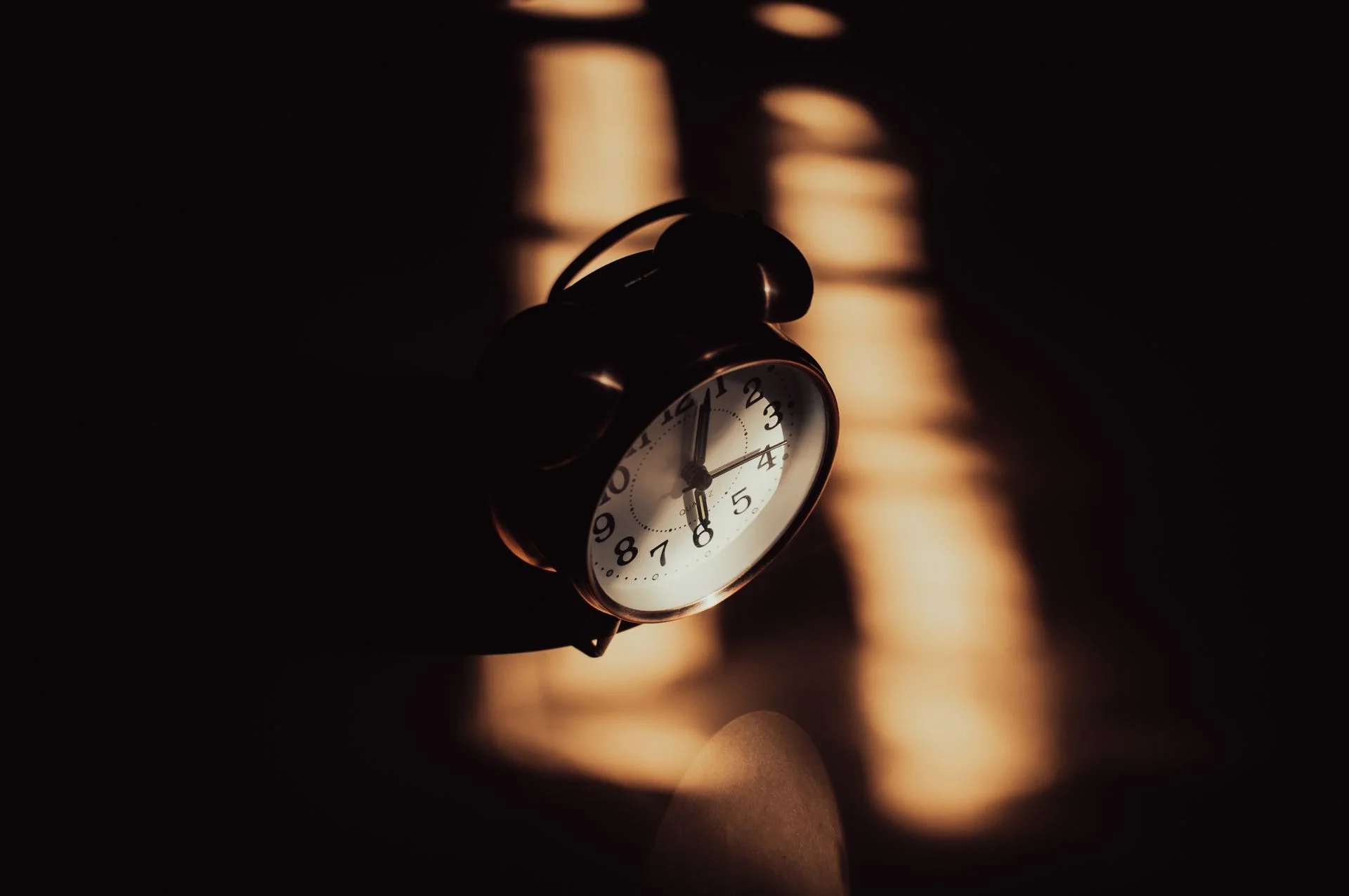 An old alarm clock sitting in a dark room, its face barely visible in the gloom — a mechanism still, even in the dark, still keeping whatever time it keeps