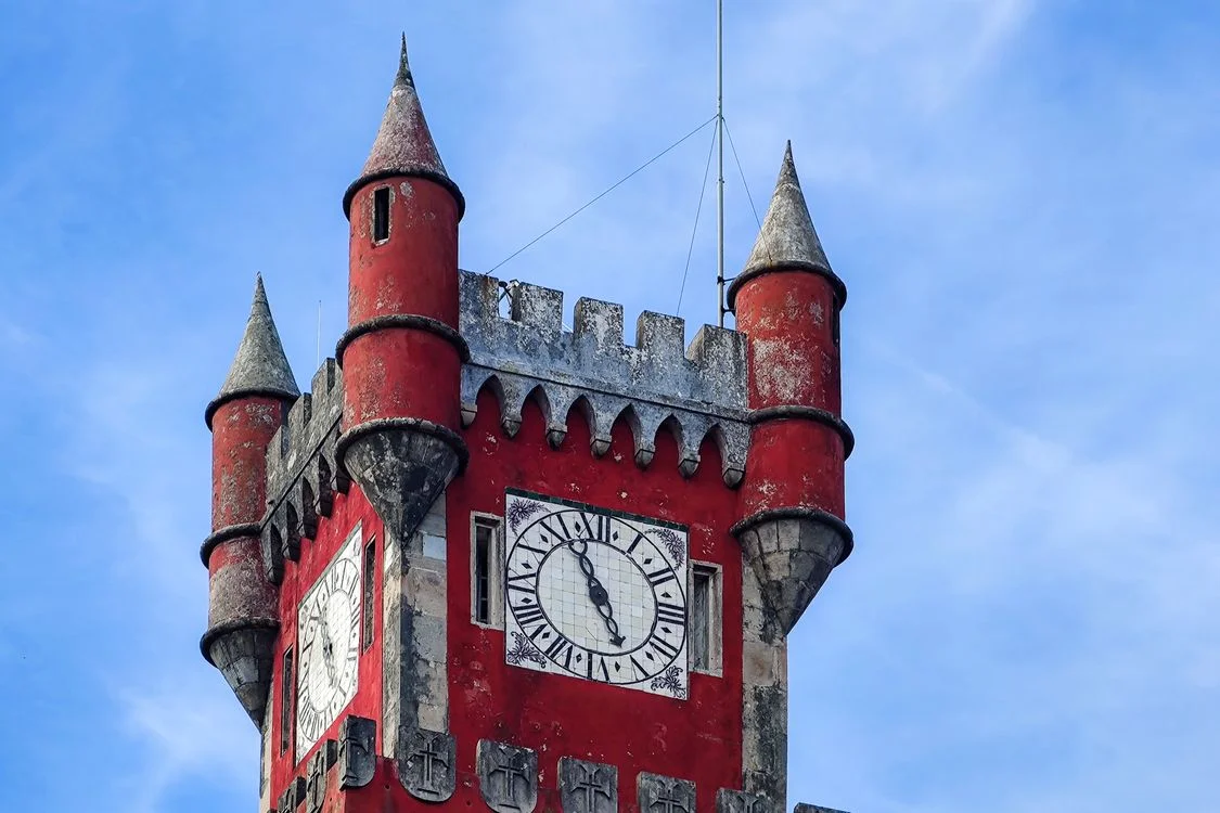 A historic castle clock tower rising against a vivid blue summer sky, the hands frozen at a moment between morning and afternoon — a structure built to mark time that now marks, for me, something more personal