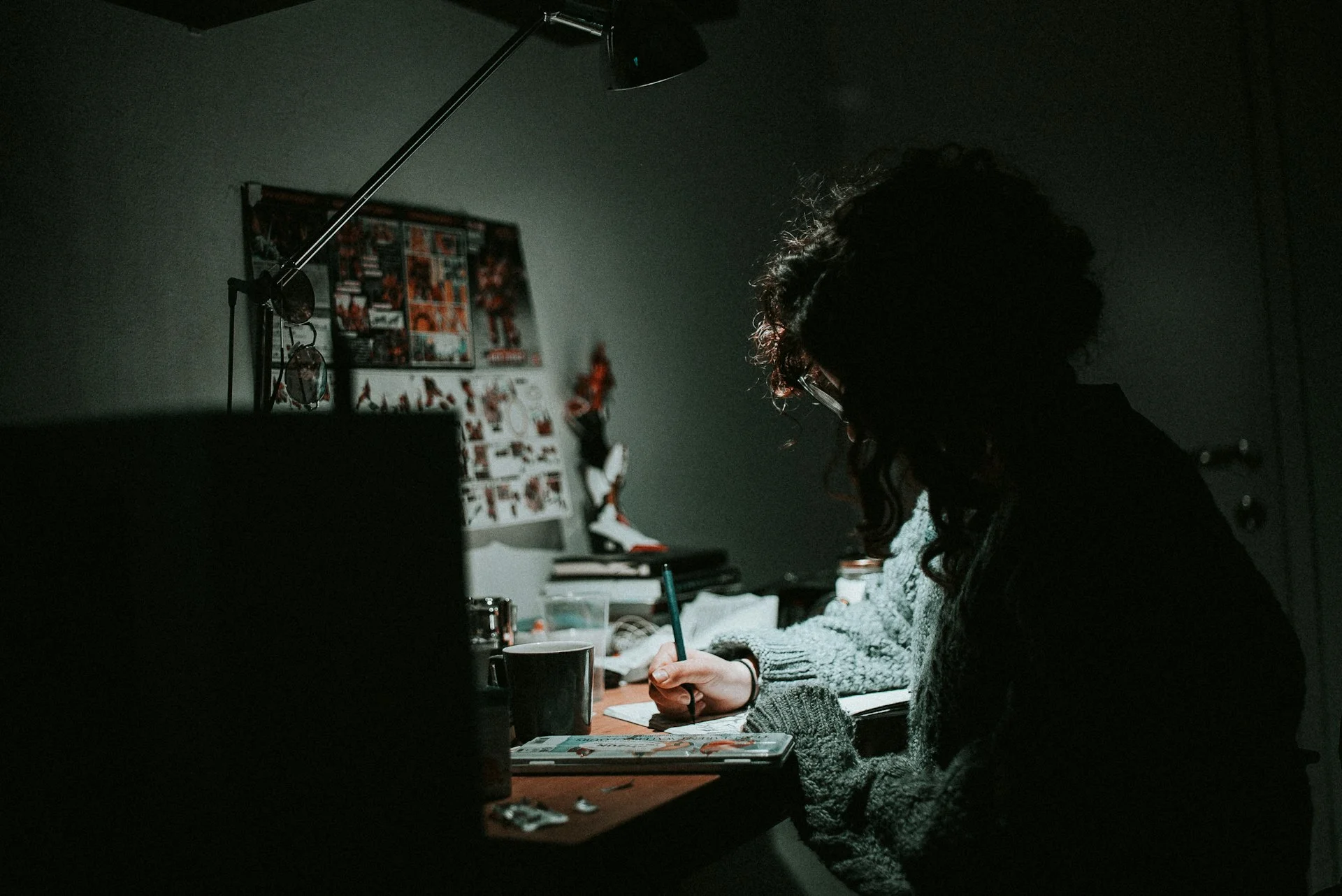 A solitary figure working late at a wide desk in warm lamplight — the room around them fading into deep shadow, the lamp the only island of warmth in a vast darkness